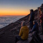 Travelers in warm layers watch sunrise from a Mt. Fuji mountain hut terrace above the clouds