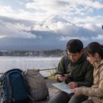 Travelers checking weather and route information beside Lake Kawaguchiko with Mt. Fuji behind clouds
