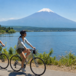 Two travelers cycling beside Lake Yamanaka with Mount Fuji across the water