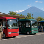 Three unbranded compact sightseeing buses in red green and blue with travelers boarding below snow-free Mount Fuji