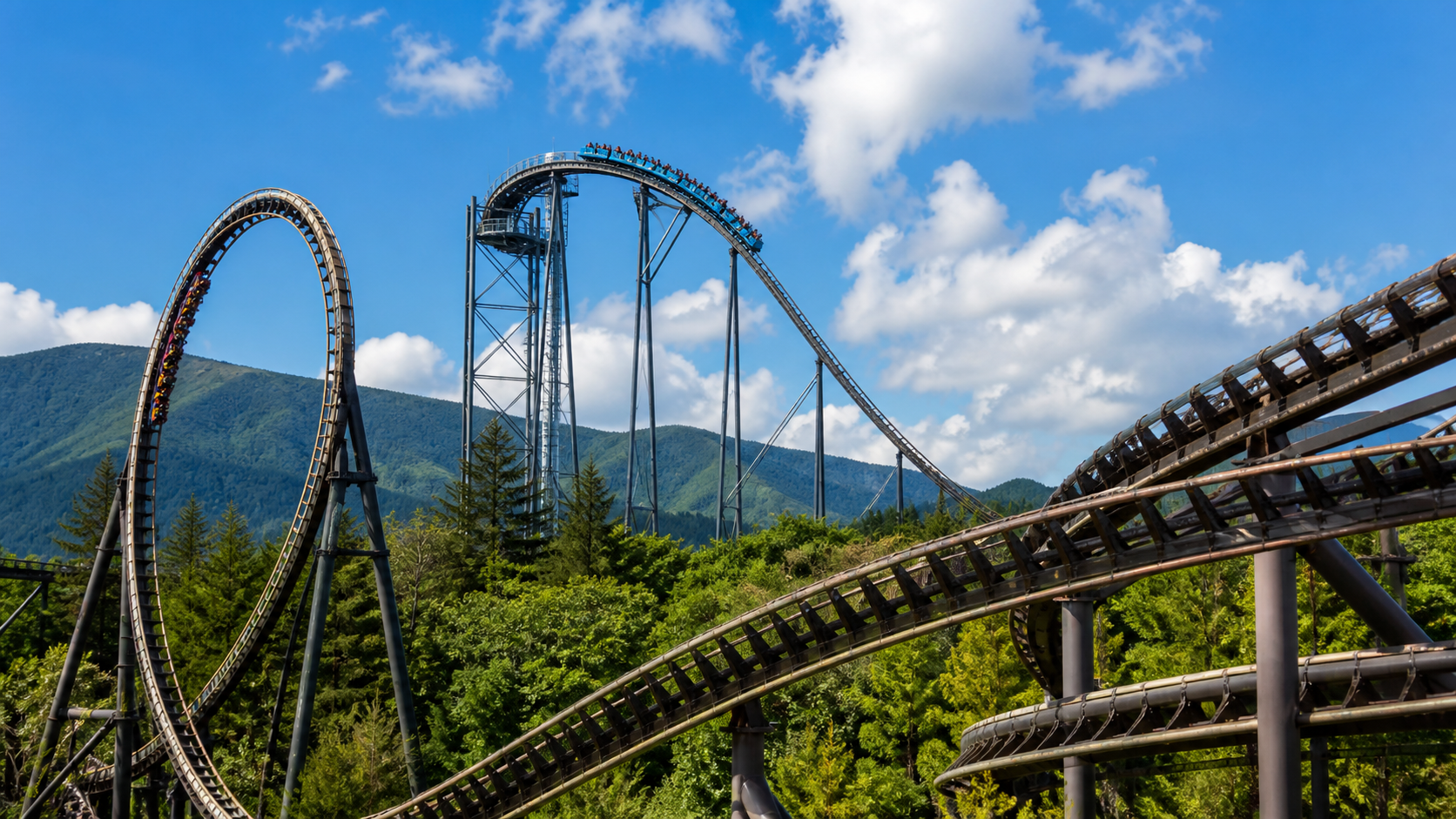 Three different steel roller coaster tracks in a mountain amusement park under a summer sky