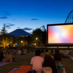 Travelers seated on a lawn facing a blank outdoor movie screen with roller-coaster tracks and snow-free Mount Fuji at dusk