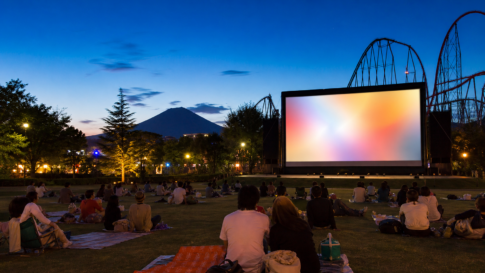 Travelers seated on a lawn facing a blank outdoor movie screen with roller-coaster tracks and snow-free Mount Fuji at dusk
