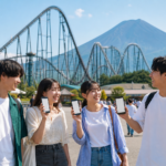 Text-free group of university-age friends holding blank smartphone tickets in front of a roller coaster and snow-free Mount Fuji