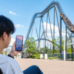 A visitor checking an unlabeled progress ring on a smartphone beside a roller coaster