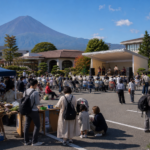 Text-free community market and small performance stage in a civic parking area near snow-free Mount Fuji