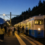 Concert visitors boarding an unbranded blue and white local train at a Mount Fuji foothills station after sunset