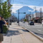 Traveler photographing snow-free Mount Fuji from a wide sidewalk while cars remain unobstructed on a Fujiyoshida street