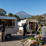 Travelers visiting blank food trucks and local produce tables at a roadside station below a snow-free Mount Fuji