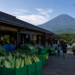 Travelers walking beside plain crates of fresh corn and vegetables at a roadside market below green Mount Fuji