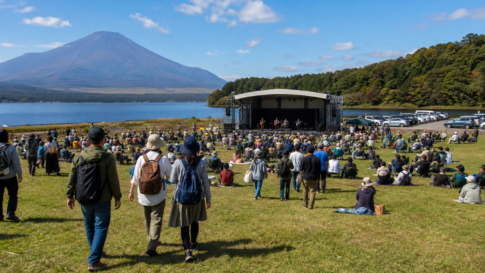 Travelers arriving at a text-free outdoor music festival beside Lake Yamanaka with bare Mount Fuji beyond