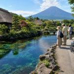 Travelers including a wheelchair user viewing a clear Oshino Hakkai spring pond from a paved path below a snow-free Mount Fuji