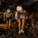 Two travelers wearing helmets and moving carefully through a low volcanic lava cave