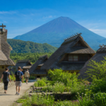Travelers walking through a restored thatched-roof village below a snow-free Mount Fuji in summer