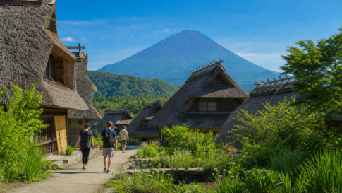 Travelers walking through a restored thatched-roof village below a snow-free Mount Fuji in summer