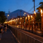 Travelers walking behind barriers beside a closed Fujiyoshida street lined with tall burning festival torches