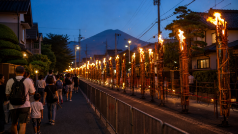 Travelers walking behind barriers beside a closed Fujiyoshida street lined with tall burning festival torches
