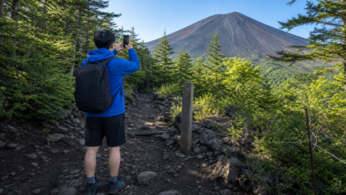 A hiker with plain unbranded gear taking a phone photo beside an unmarked wooden checkpoint on the summer Yoshida Trail